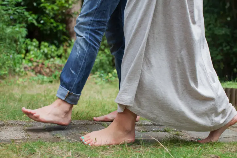 View of a man and a woman from the knees down. They are walking along a path, both barefoot. The man is wearing jeans, the woman a flowing light grey skirt.